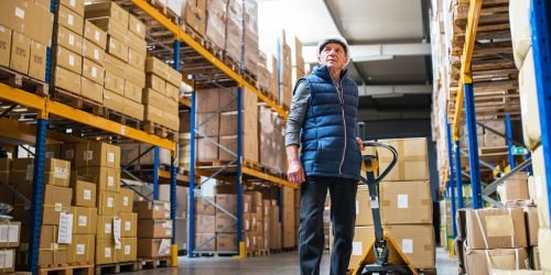 Senior male warehouse worker pulling a pallet truck with boxes.